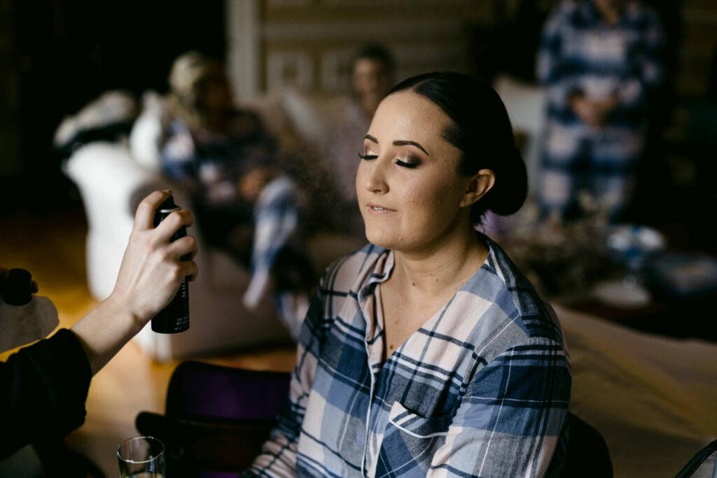 Bride getting her makeup set with a fine mist of setting spray on the morning of her wedding at Buckland Estate, wearing a blue plaid shirt with bridesmaids relaxing in the background.