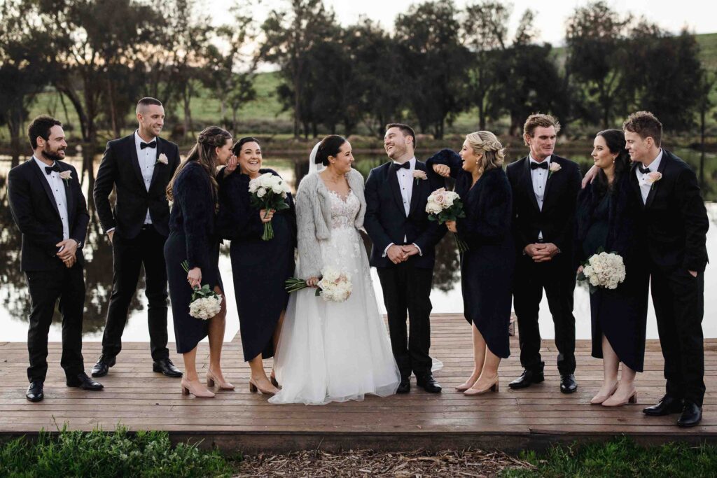 Bride and groom laughing with their wedding party on a wooden dock at Buckland Estate, holding bouquets of white and blush flowers.