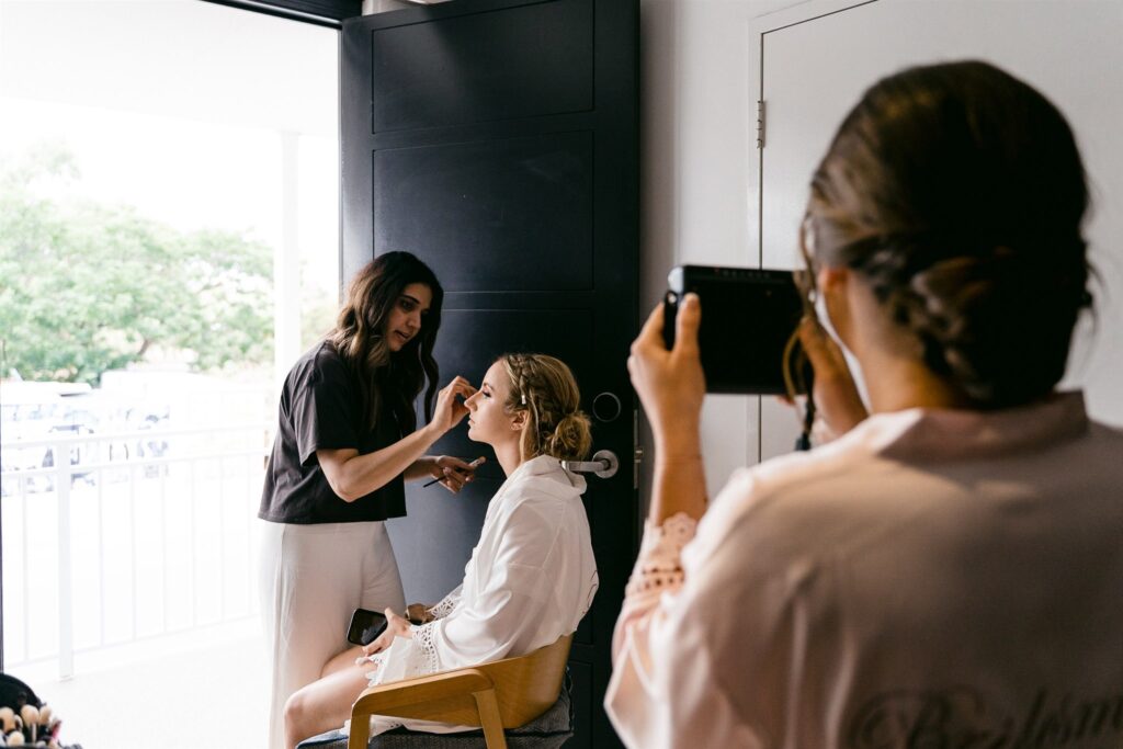 Bride getting her makeup done while a bridesmaid captures the moment — a key wedding photo preparation tip.