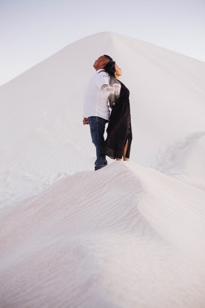 Couple standing back-to-back in Lancelin dunes as sand pours from their hands, showcasing one of the top Western Australia pre-wedding photo locations.