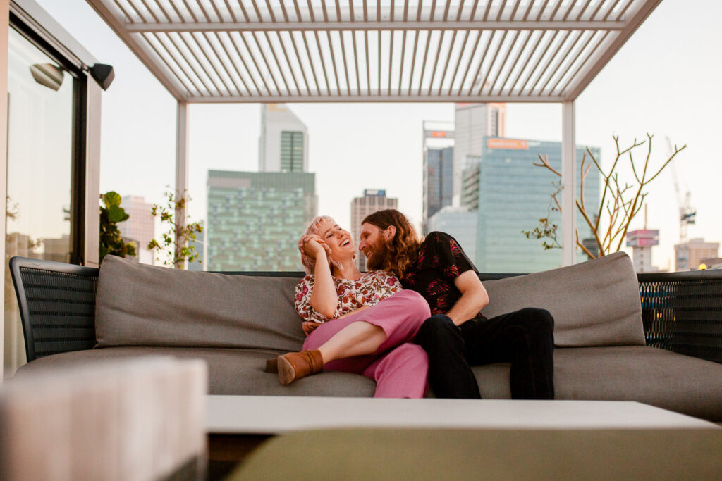 Couple laughing on a rooftop lounge during a pre-wedding shoot in Perth with city skyline behind them.