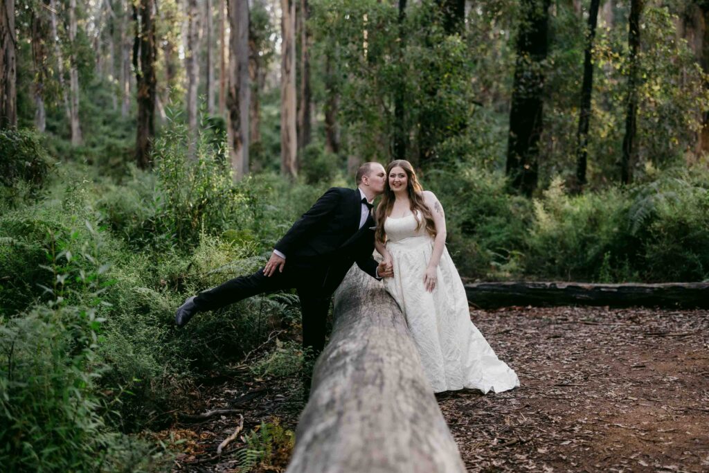 husband leaning over log to kiss bride on the cheek in boranup forrrest for pre-wedding photos